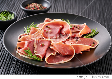 parma ham prosciutto with rosemary and spices in a plate on a black wooden table. parma ham prosciutto with rosemary and spices in a plate on a black wooden table. 109794296