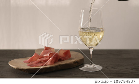 pour white whine in wine glass on oak table with bresaola on olive board pour white whine in wine glass on oak table with bresaola on olive board 109794837