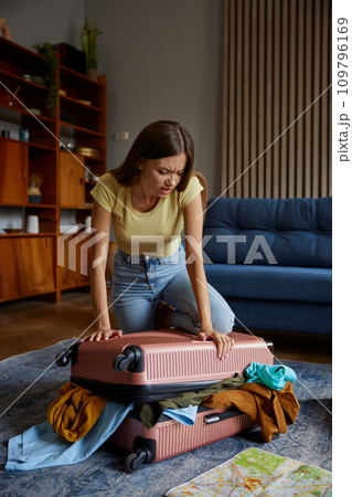 Young woman struggling with overload travel suitcase while preparing for trip Young woman struggling with overload travel suitcase while preparing for trip 109796169