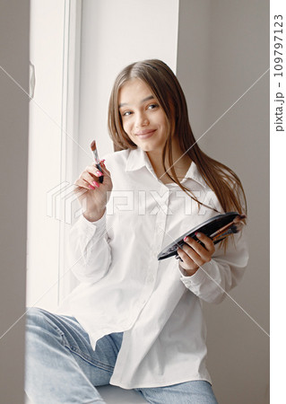 Young female artist posing holding brushes for make up and palette in hands. Woman wearing white shirt. Brunette girl sitting on a windowsill in room with white wall. Young female artist posing holding brushes for make up and palette in hands. Woman wearing white shirt. Brunette girl sitting on a windowsill in room with white wall. 109797123