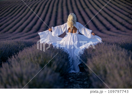 Blonde woman poses in lavender field at sunset. Happy woman in white dress holds lavender bouquet. Aromatherapy concept, lavender oil, photo session in lavender 109797743