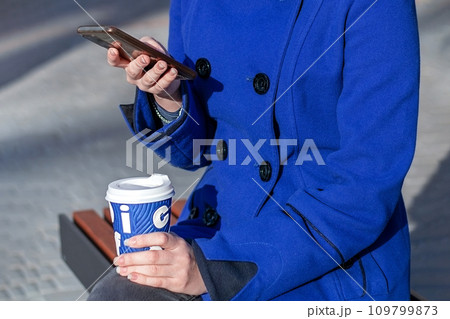 A girl looks at her phone and drinks coffee while sitting on a bench on the street. Without a face A girl looks at her phone and drinks coffee while sitting on a bench on the street. Without a face 109799873