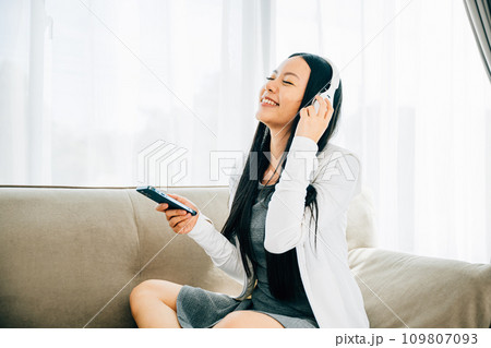 Attractive woman on sofa listens to music uses smartphone with headphones. Engaged in leisure relaxation and entertainment at home. Modern technology concept. 109807093