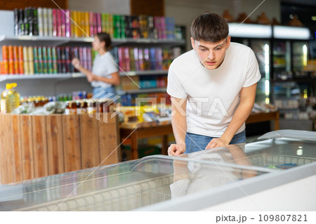 Young guy looks and chooses frozen food in refrigerator in supermarket 109807821