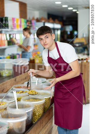 Friendly salesman in an apron offers to buy pickled green olives in grocery supermarket Friendly salesman in an apron offers to buy pickled green olives in grocery supermarket 109807830