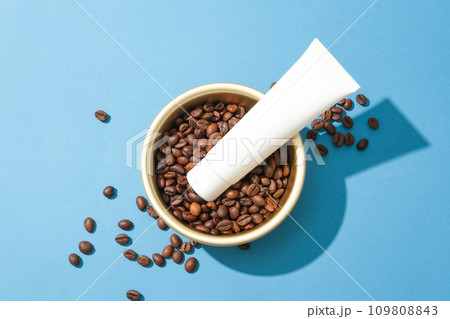 Close-up of a cosmetic tube placed on top of coffee beans in a ceramic bowl. Blue background for advertising. Coffee has antibacterial properties that help limit acne on the skin. Close-up of a cosmetic tube placed on top of coffee beans in a ceramic bowl. Blue background for advertising. Coffee has antibacterial properties that help limit acne on the skin. 109808843