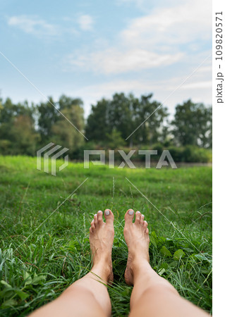 The image shows a pair of bare feet lying on a grassy field with a lake and a green park in the background. Toes point upward. The image is peaceful and calm. 109820571