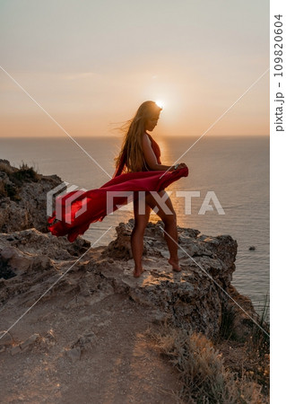 Woman sunset sea red dress, side view a happy beautiful sensual woman in a red long dress posing on a rock high above the sea on sunset. 109820604