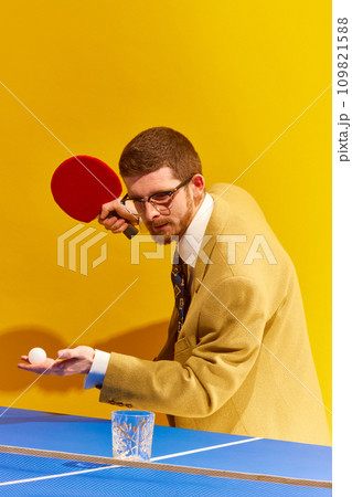 Concentrated young man in glasses, elegant suit playing table tennis, hitting ball into whiskey glass against bright yellow background 109821588