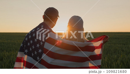 Happy couple with american flag on their shoulders looking forward in wheat field at sunset 109821680
