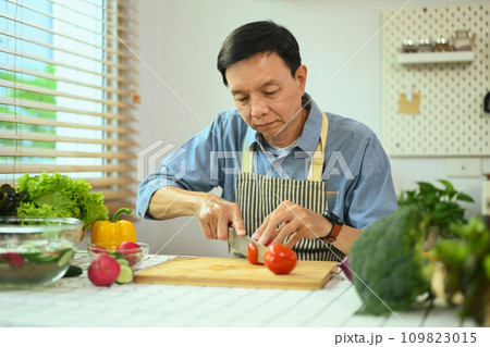 Middle age Asian man slicing tomatoes over a table for a vegan meal. Healthy lifestyle concept. Middle age Asian man slicing tomatoes over a table for a vegan meal. Healthy lifestyle concept. 109823015