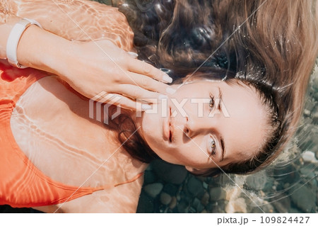 Woman travel portrait. close-up portrait of a happy woman with long hair in a red bikini, floating in water and smiling at the camera. 109824427