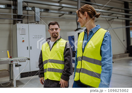 Young man with Down syndrome working in warehouse, colleague talking with him. Concept of workers with disabilities, support in workplace. Young man with Down syndrome working in warehouse, colleague talking with him. Concept of workers with disabilities, support in workplace. 109825002