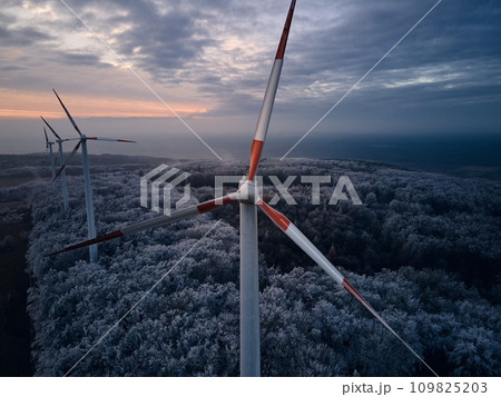 Aerial landscape photography of sunrise over frost-covered nature with wind turbines. Windmill in soft morning light with icy trees around. Concept of wind power as clean, renewable energy source. Aerial landscape photography of sunrise over frost-covered nature with wind turbines. Windmill in soft morning light with icy trees around. Concept of wind power as clean, renewable energy source. 109825203