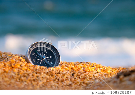 Conceptual photo, close-up of a compass lying in wet sand consisting of small crumbs of shells, against the backdrop of a blue calm sea Conceptual photo, close-up of a compass lying in wet sand consisting of small crumbs of shells, against the backdrop of a blue calm sea 109829107