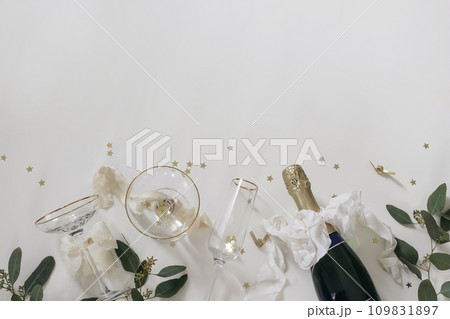 New year party, celebration banner. Bottle of champagne wine, drinking glasses, bows. Eucalyptus tree branches isolated on white table background. Golden star confetti. Birthday flat lay, top view. New year party, celebration banner. Bottle of champagne wine, drinking glasses, bows. Eucalyptus tree branches isolated on white table background. Golden star confetti. Birthday flat lay, top view. 109831897