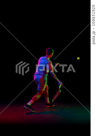 Dynamic image of man, athlete in motion, playing tennis, serving ball with racket against dark background in neon light Dynamic image of man, athlete in motion, playing tennis, serving ball with racket against dark background in neon light 109831920