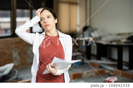 Confused female designer standing with papers in apartment during renovation 109842338