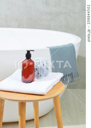 A bottle of brown shampoo and a bath sponge on a folded towel on a wooden table, next to a bathtub on a gray background. Space inside the bathroom for advertising. 109844501