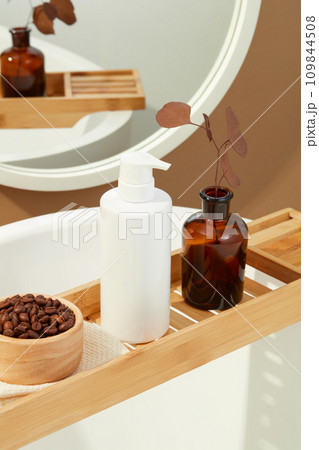 Coffee beans in a wooden bowl, a bottle of shower gel without a label, a brown vase are displayed on a wooden tray. Space inside the bathroom with bathtub, mirror and body care products. Coffee beans in a wooden bowl, a bottle of shower gel without a label, a brown vase are displayed on a wooden tray. Space inside the bathroom with bathtub, mirror and body care products. 109844508