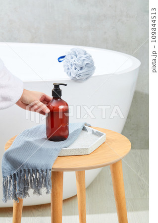 A hand holds an unlabeled bottle of shampoo placed on a tray with a cloth towel on a wooden table, behind which is a bathtub and a shower sponge. Minimalist gray background for advertising. A hand holds an unlabeled bottle of shampoo placed on a tray with a cloth towel on a wooden table, behind which is a bathtub and a shower sponge. Minimalist gray background for advertising. 109844513