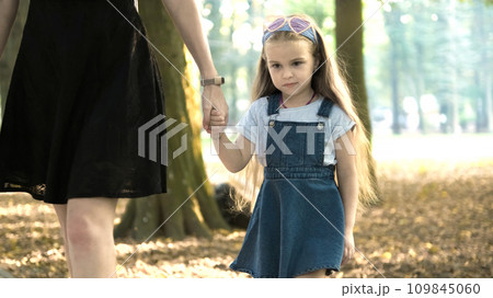 Mom and her small daughter with long hair walking together holding hands in summer park. 109845060