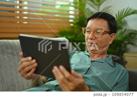 Carefree senior man browsing internet on digital tablet while relaxing on a couch at home. 109854077