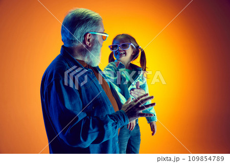 Senior man and little girl, grandfather and granddaughter watching TV over gradient orange background in neon light Senior man and little girl, grandfather and granddaughter watching TV over gradient orange background in neon light 109854789