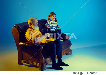 Senior man and little girl, grandfather watching TV with his granddaughter over blue background in neon light 109854808