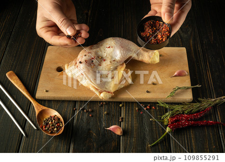 Raw broiler leg on a kitchen cutting board. Adding pepper by hand and spoon to a chicken leg before baking in the oven 109855291