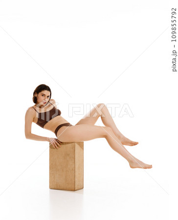 Captivating full length portrait of featuring young woman in brown underwear sitting on wood cube against pristine white background. Captivating full length portrait of featuring young woman in brown underwear sitting on wood cube against pristine white background. 109855792