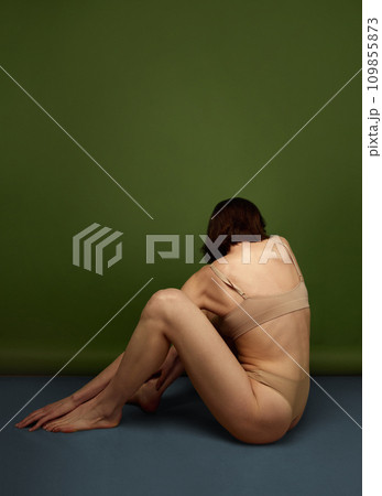 Side view portrait of brunette woman posing sitting on floor in flesh color lingerie against olive studio background. 109855873