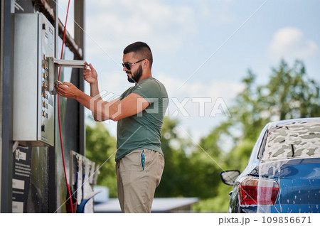 Young man paying for cleaning car on self-service car wash. Cropped image of blue auto and man that going to wash it by active foam. Male person paying for wash service next to car at sunny day. 109856671