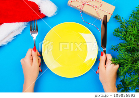 Hungry woman waiting for meal. empty yellow plate on a blue background with Christmas branches, santa hat and gift box. perspective view for your design Hungry woman waiting for meal. empty yellow plate on a blue background with Christmas branches, santa hat and gift box. perspective view for your design 109856992