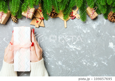 Top view of a woman holding a gift box in her hands on festive cement background. Fir tree and Christmas decorations. New year time concept Top view of a woman holding a gift box in her hands on festive cement background. Fir tree and Christmas decorations. New year time concept 109857695