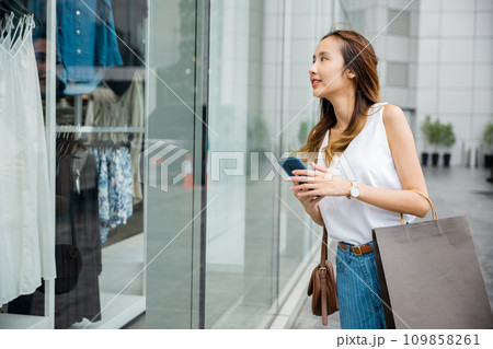 Happy woman with bags and a smartphone, standing outside a fashion store on a sunny day. She is enjoying the convenience of technology and the pleasure of shopping. Happy woman with bags and a smartphone, standing outside a fashion store on a sunny day. She is enjoying the convenience of technology and the pleasure of shopping. 109858261