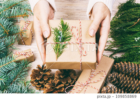 Hand crafted gift on wooden background with Christmas decor. Woman wrapping christmas present , girl prepares xmas gifts with fir tree and pine cone. Top view, copy space. Toned 109858869