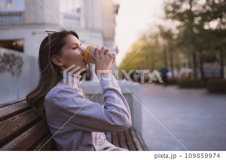 Woman drinks from cup on wooden bench. She is wearing a white shirt enjoying her beverage. The bench is located in a park setting, with trees in the background. Woman drinks from cup on wooden bench. She is wearing a white shirt enjoying her beverage. The bench is located in a park setting, with trees in the background. 109859974