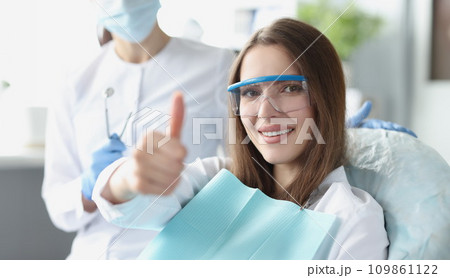 Young woman in safety glasses sitting in dental chair and showing thumbs up 109861122