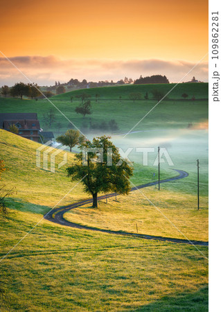 Rural scene with curved road, tree and small village in the valley and foggy morning at Switzerland Rural scene with curved road, tree and small village in the valley and foggy morning at Switzerland 109862281
