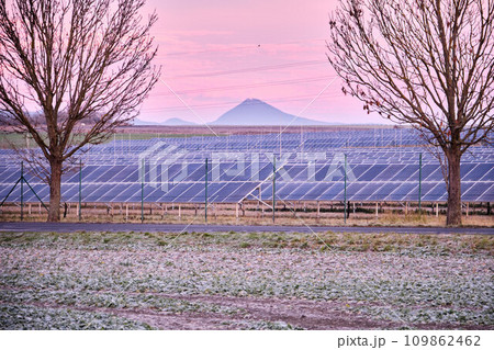 Solar power plant park with panels beyond chain link fence. Subsidized renewable energy source on arable land. Europe. Czech Republic. Freezing winter morning or evening in agricultural field. 109862462