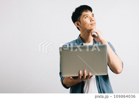 An Asian engineer, deep in thought, holds a laptop, pondering questions and seeking answers with a focused expression. Studio shot isolated on white, where success is born from inquiry. An Asian engineer, deep in thought, holds a laptop, pondering questions and seeking answers with a focused expression. Studio shot isolated on white, where success is born from inquiry. 109864036