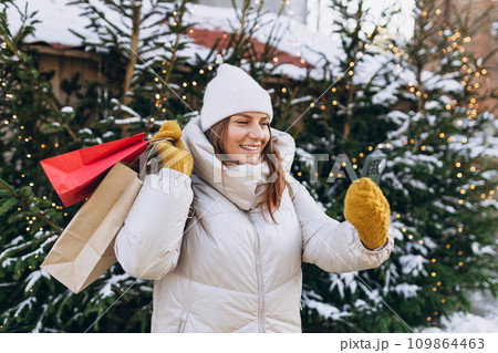 Happy cheerful Woman with backpack holding red shopping paper bags in hands, walking at street. Young hipster with presents taking selfie. Winter holidays sale. Shopping banner Happy cheerful Woman with backpack holding red shopping paper bags in hands, walking at street. Young hipster with presents taking selfie. Winter holidays sale. Shopping banner 109864463
