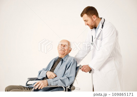 Senior gentleman on a wheelchair isolated on a white background. Man wearing blue t-shirt and doctor wearing white medical gown. Doctor talking with man. Senior gentleman on a wheelchair isolated on a white background. Man wearing blue t-shirt and doctor wearing white medical gown. Doctor talking with man. 109865171