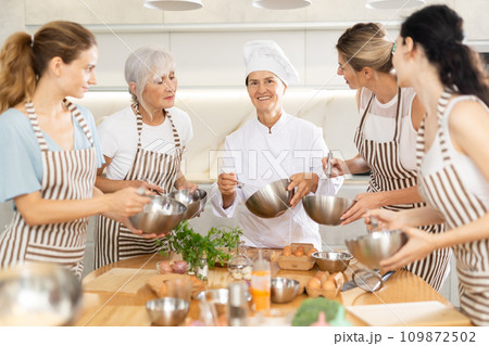 Elderly woman chef with bowl in hands tells women at culinary courses about traditions of cuisine Elderly woman chef with bowl in hands tells women at culinary courses about traditions of cuisine 109872502