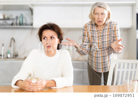 Upset middle-aged woman sitting at the kitchen table with her back to old woman quarreling to her 109872951