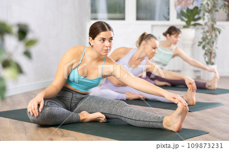 Diligent women practicing hand to toe pose of yoga in light fitness room Diligent women practicing hand to toe pose of yoga in light fitness room 109873231