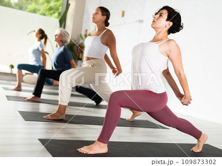 Focused young Latin woman balancing in Virabhadrasana 1 Warrior pose on mats during group yoga class Focused young Latin woman balancing in Virabhadrasana 1 Warrior pose on mats during group yoga class 109873302