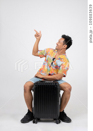 A cheerful Asian man tourist is sitting on his luggage and pointing his finger at an empty space. 109883639