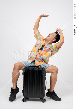 Excited, fun Asian man in summer clothes is sitting on his luggage on an isolated white background. 109883643